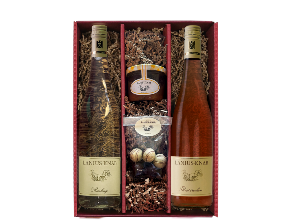 roter Geschenkkarton mit einer hellen Weinflasche gefüllt mit Weißwein, einer hellen Weinflasche gefüllt mit Rosé, einem Glas Chutney und einer Tüte mit weißen und dunklen Pralinen